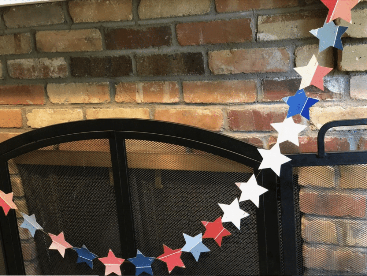A Fourth of July star-shaped garland in patriotic colors draped on a fireplace screen against a brick backdrop.