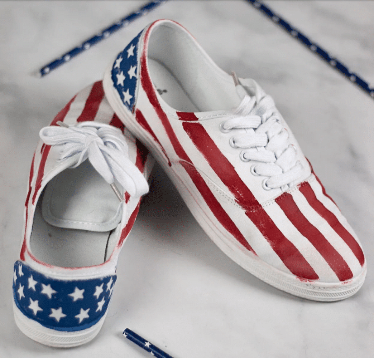 A pair of sneakers painted with the American flag design, featuring stripes and stars, on a white surface next to red and blue pencils for Fourth of July crafts.