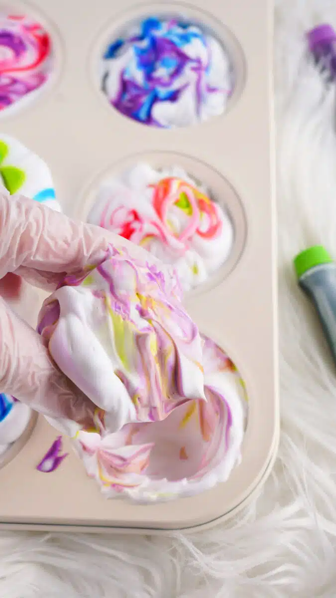Hand in a glove lifting a cupcake with swirling colorful icing, resembling shaving cream, from a muffin tin.