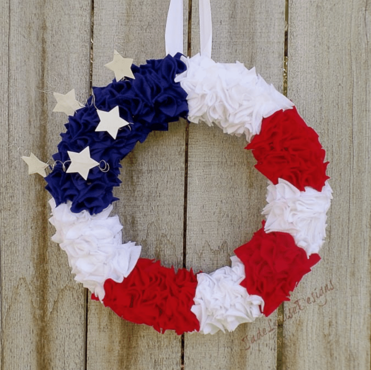 A patriotic Fourth of July wreath featuring red, white, and blue fabric flowers with white stars against a wooden backdrop.