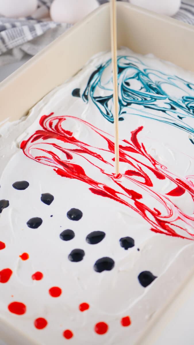 Marbled paint design being swirled with a wooden stick on a white background with red, blue, and black paint drops on Cool Whip eggs.