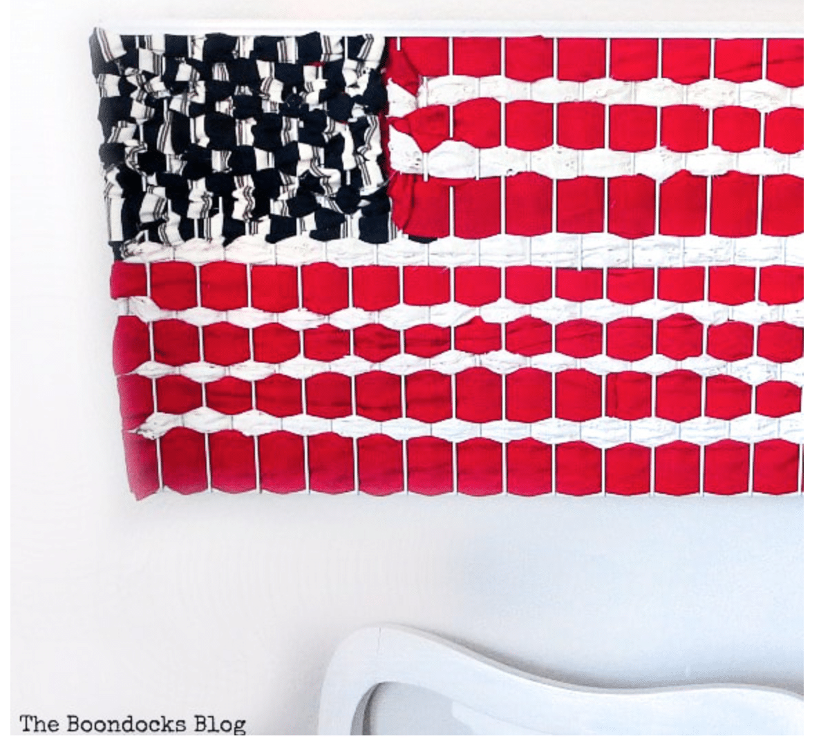 A handmade wall hanging with a Fourth of July crafts pattern resembling the American flag displayed above a white chair.