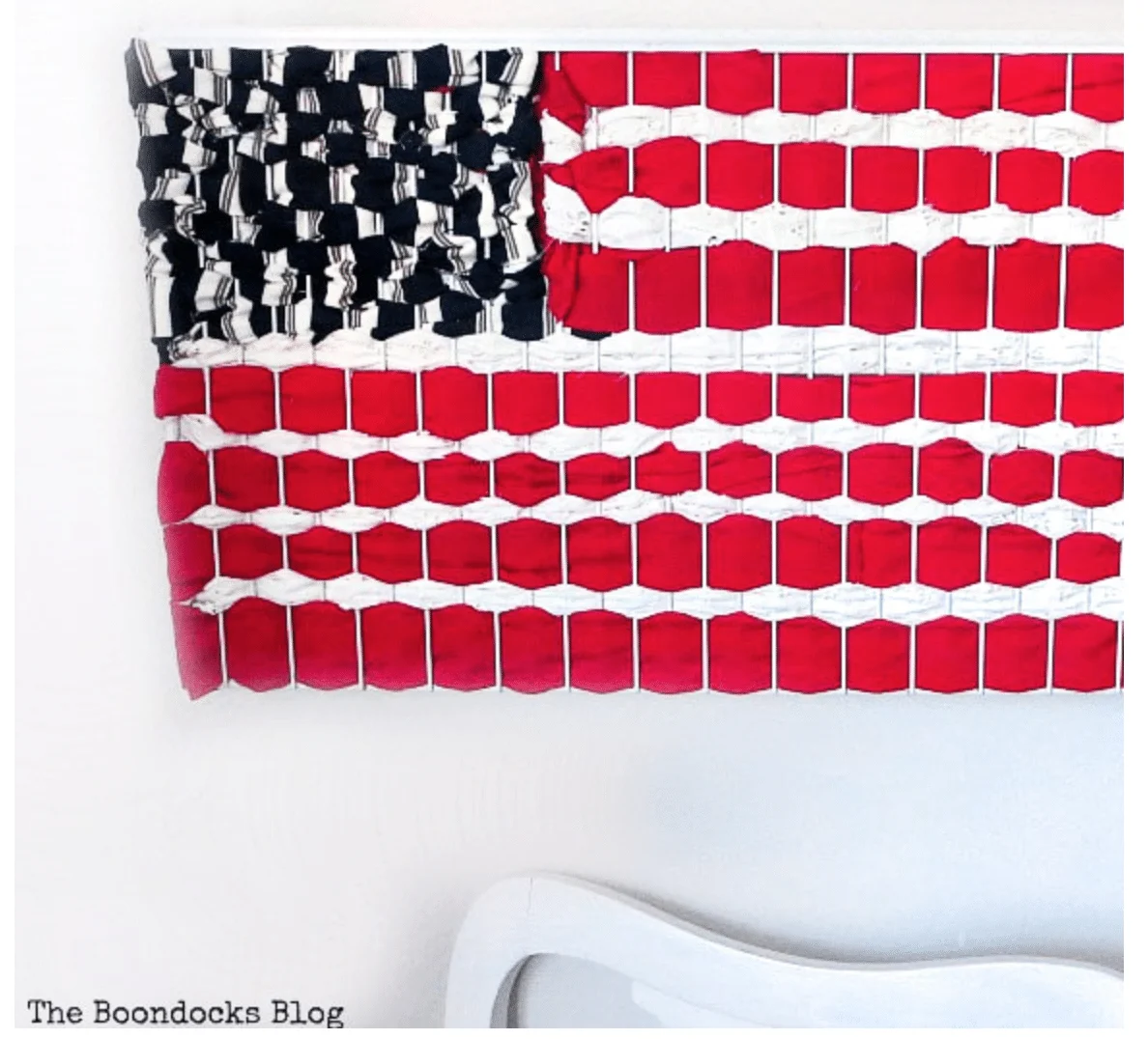 A handmade wall hanging with a Fourth of July crafts pattern resembling the American flag displayed above a white chair.