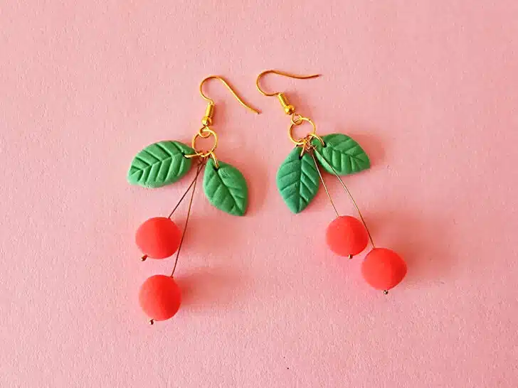 A pair of cherry-themed earrings with gold hooks, green leaf accents, and red cherry beads against a pink background, these delightful clay cherry earrings add a touch of whimsy.