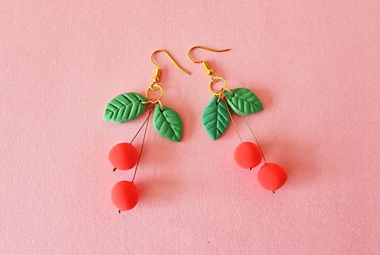 A pair of cherry-themed earrings with gold hooks, green leaf accents, and red cherry beads against a pink background, these delightful clay cherry earrings add a touch of whimsy.