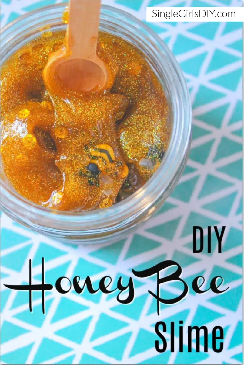 A jar of glittery, honey-colored DIY slime with a small wooden spoon on top of a blue and white patterned surface. The text on the image reads "DIY Honey Bee Slime.
