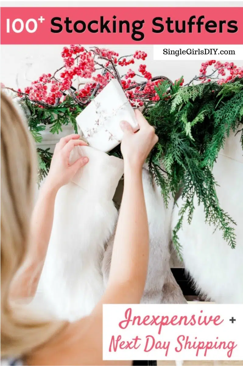 Person placing a wrapped gift into a white stocking adorned with holiday greenery and red berries. Text on the image reads "100+ Inexpensive Stocking Stuffers," "Inexpensive + Next Day Shipping," and "SingleGirlsDIY.com.