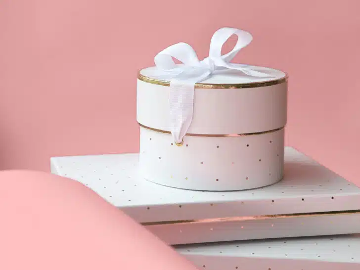 A round white gift box with a white ribbon sits gracefully on top of white polka-dotted rectangular boxes against a pink background, perfect for your gift closet.