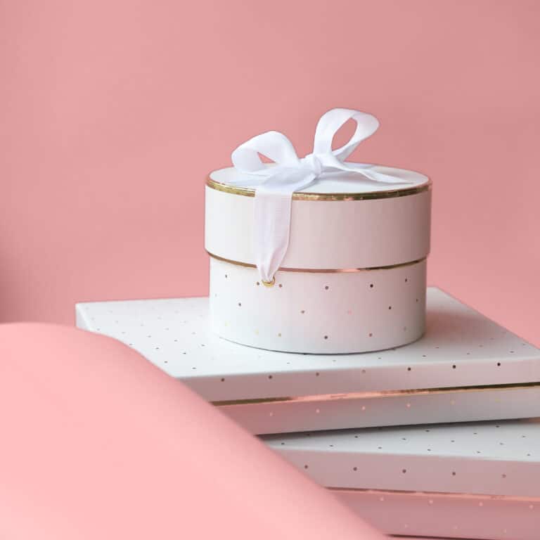 A round white gift box with a white ribbon sits gracefully on top of white polka-dotted rectangular boxes against a pink background, perfect for your gift closet.