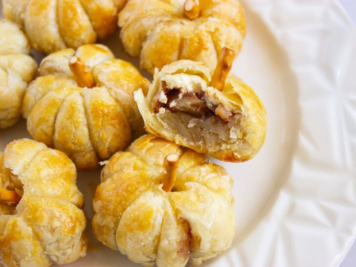 A plate of puff pumpkin pastries with pretzel sticks as stems; one pastry is bitten, revealing a filling of sliced apples and spices.