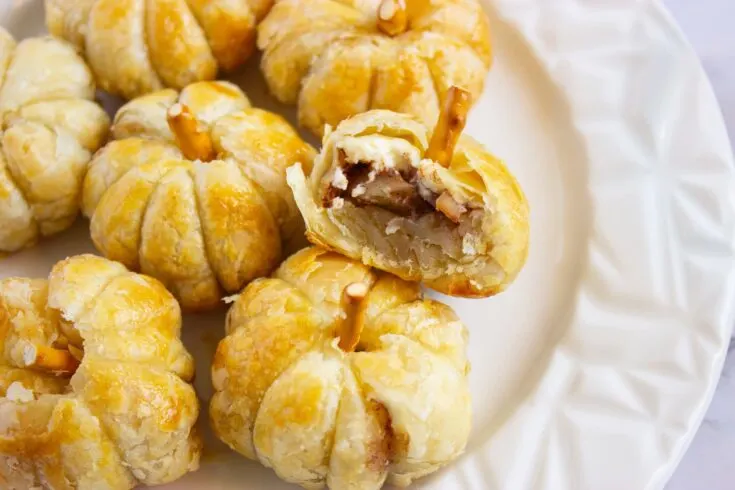 A plate of puff pumpkin pastries with pretzel sticks as stems; one pastry is bitten, revealing a filling of sliced apples and spices.