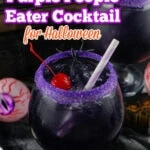 A dark purple people eater cocktail in a glass with a purple sugar rim, ice, a cherry, and a striped straw, styled for Halloween with spooky decorations in the background.