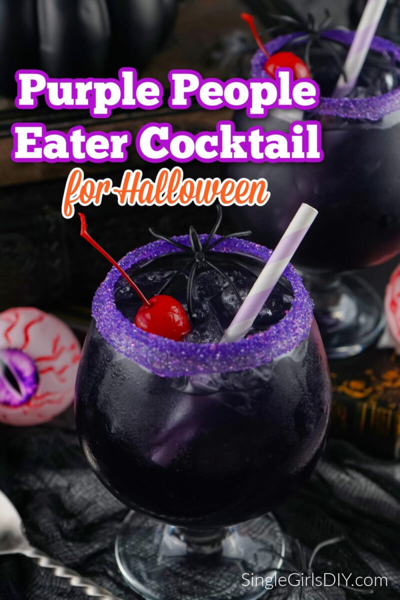 A dark purple people eater cocktail in a glass with a purple sugar rim, ice, a cherry, and a striped straw, styled for Halloween with spooky decorations in the background.