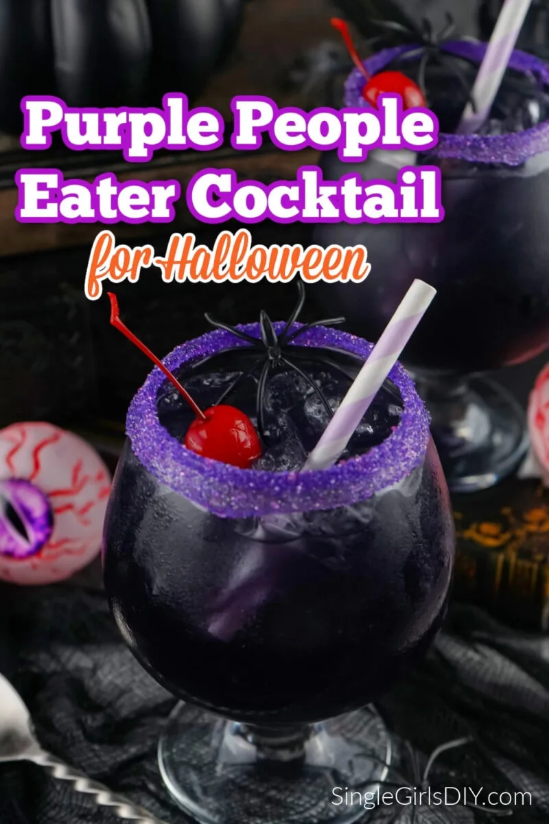 A dark purple people eater cocktail in a glass with a purple sugar rim, ice, a cherry, and a striped straw, styled for Halloween with spooky decorations in the background.