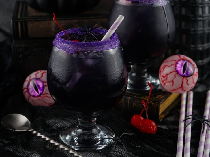 A spooky, dark purple drink with a straw and purple sugar rim, garnished with cherries, surrounded by decorative eyeballs, a silver spoon, and straws on a dark surface.