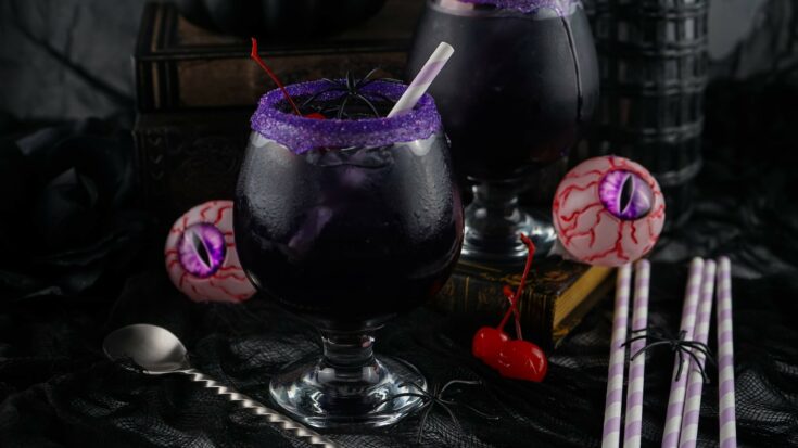 A spooky, dark purple drink with a straw and purple sugar rim, garnished with cherries, surrounded by decorative eyeballs, a silver spoon, and straws on a dark surface.