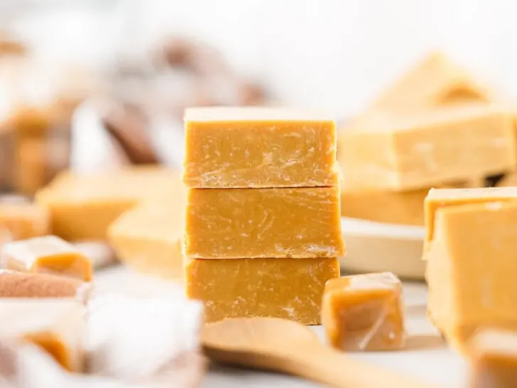 Three stacked blocks of caramel fudge on a table with more fudge pieces and a wooden spoon in the background.