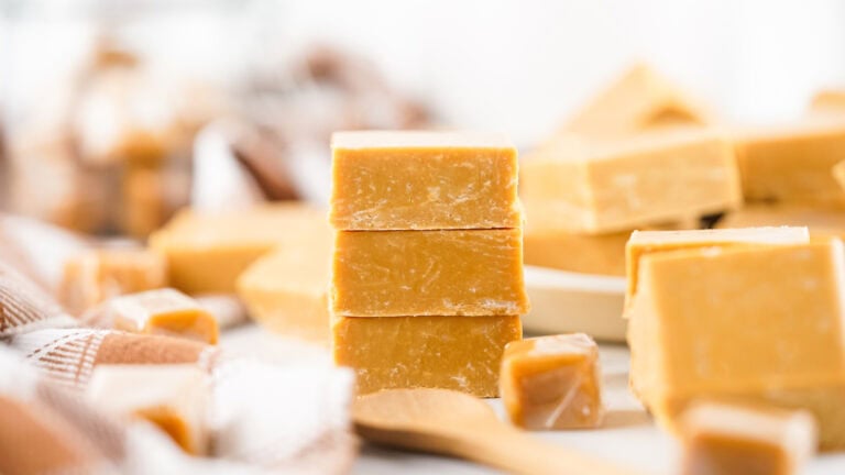 Three stacked blocks of caramel fudge on a table with more fudge pieces and a wooden spoon in the background.