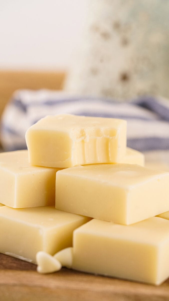 A stack of square white chocolate pieces rests on a wooden surface, with one piece showing a bite mark, reminiscent of velvety vanilla fudge.