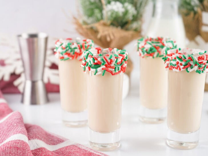 Four glasses of creamy holiday drinks topped with red, green, and white sprinkles, set on a festive table with a red cloth and snowflake decor in the background.