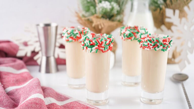 Four glasses of creamy holiday drinks topped with red, green, and white sprinkles, set on a festive table with a red cloth and snowflake decor in the background.