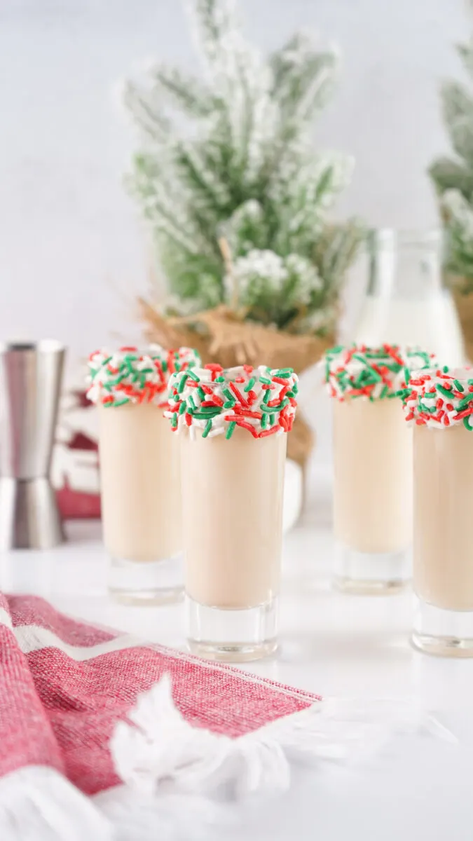 Four festive drinks in glasses with red, green, and white sprinkle rims. A red and white cloth and small potted plants are in the background.