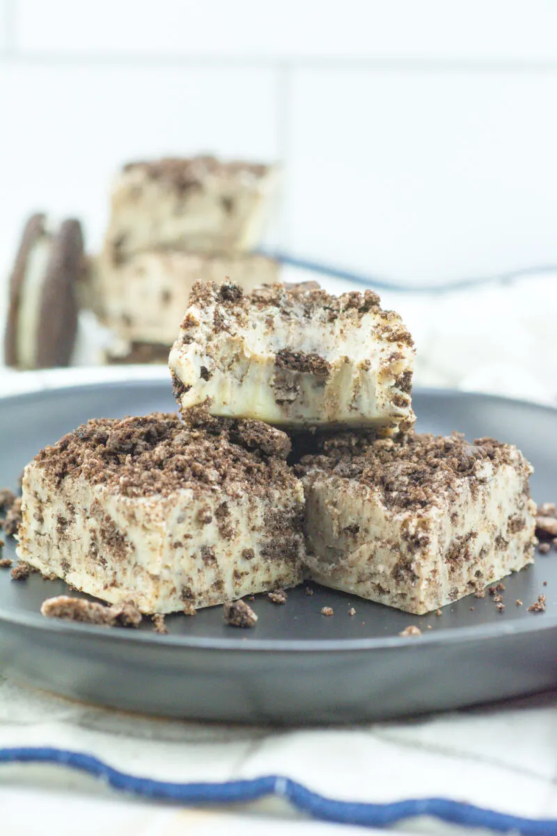 Three squares of cookies and cream fudge stacked on a dark plate, topped with crushed cookies, against a blurred background of similar treats.