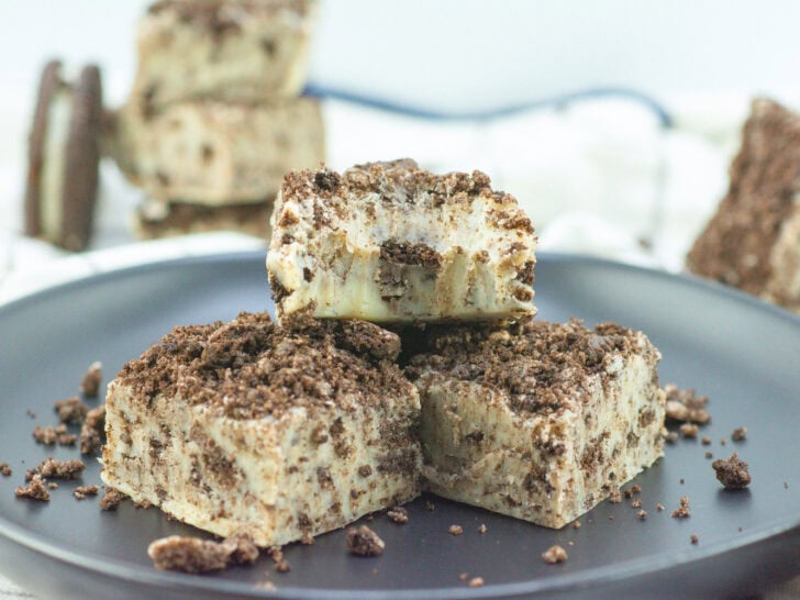 Three cookies and cream fudge squares on a black plate, with crumbled cookies on top and around them.