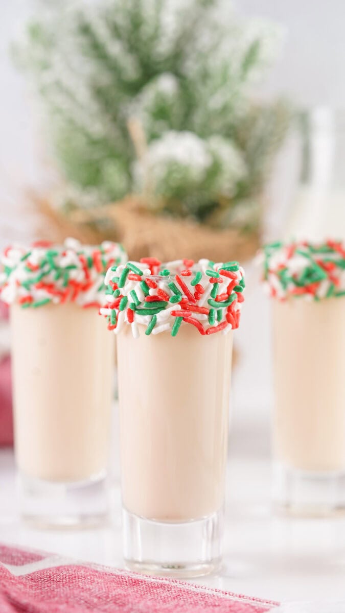 Three glasses of creamy drinks with red, white, and green sprinkle rims. Festive background with greenery and soft lighting.