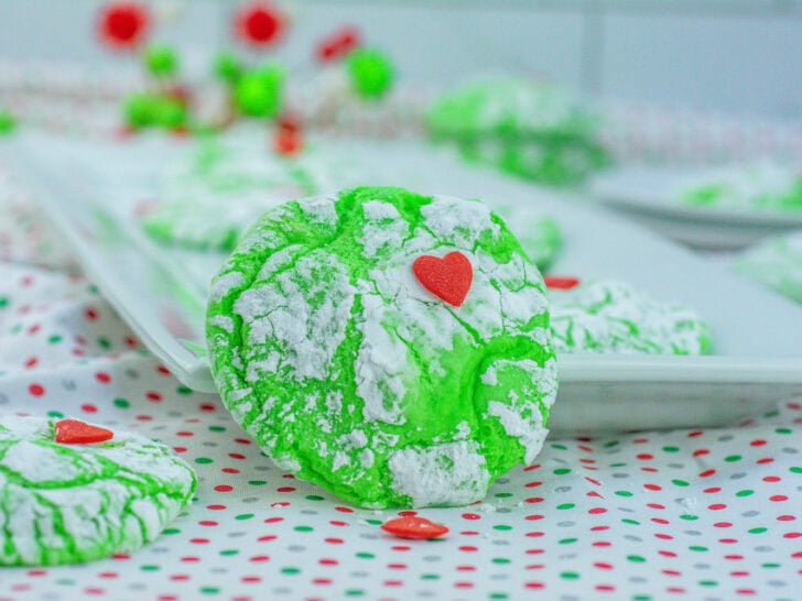 Close-up of bright green cookies with powdered sugar, each topped with a small red heart, arranged on a dotted cloth and a platter in the background.