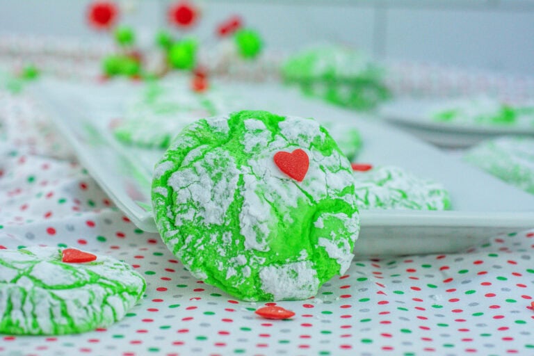 Close-up of bright green cookies with powdered sugar, each topped with a small red heart, arranged on a dotted cloth and a platter in the background.