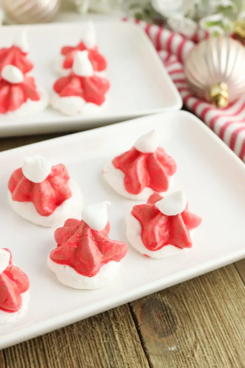 Red and white meringue cookies shaped like Santa hats are displayed on white rectangular plates, set on a wooden table next to a striped cloth and a decorative ornament.