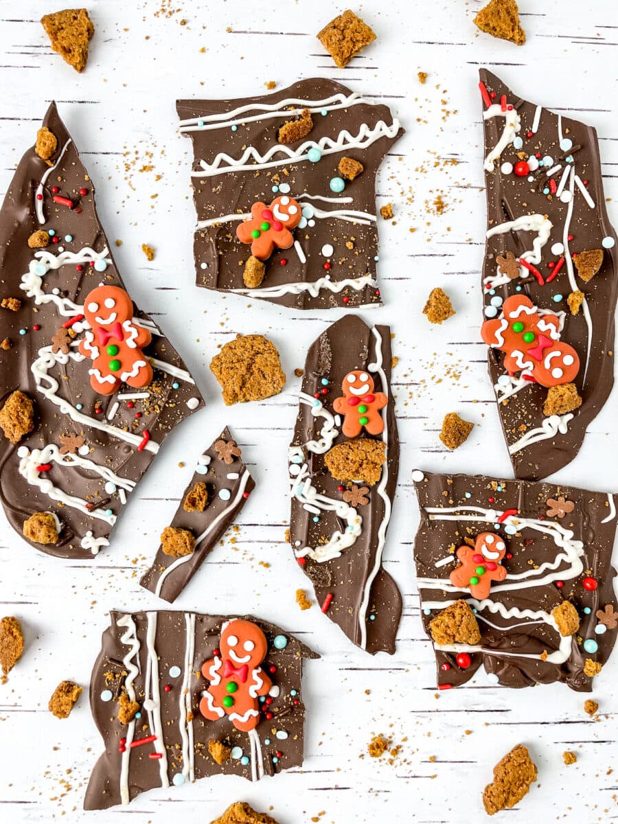 Pieces of decorated chocolate bark with gingerbread figures, icing, and colorful sprinkles scattered on a white surface.