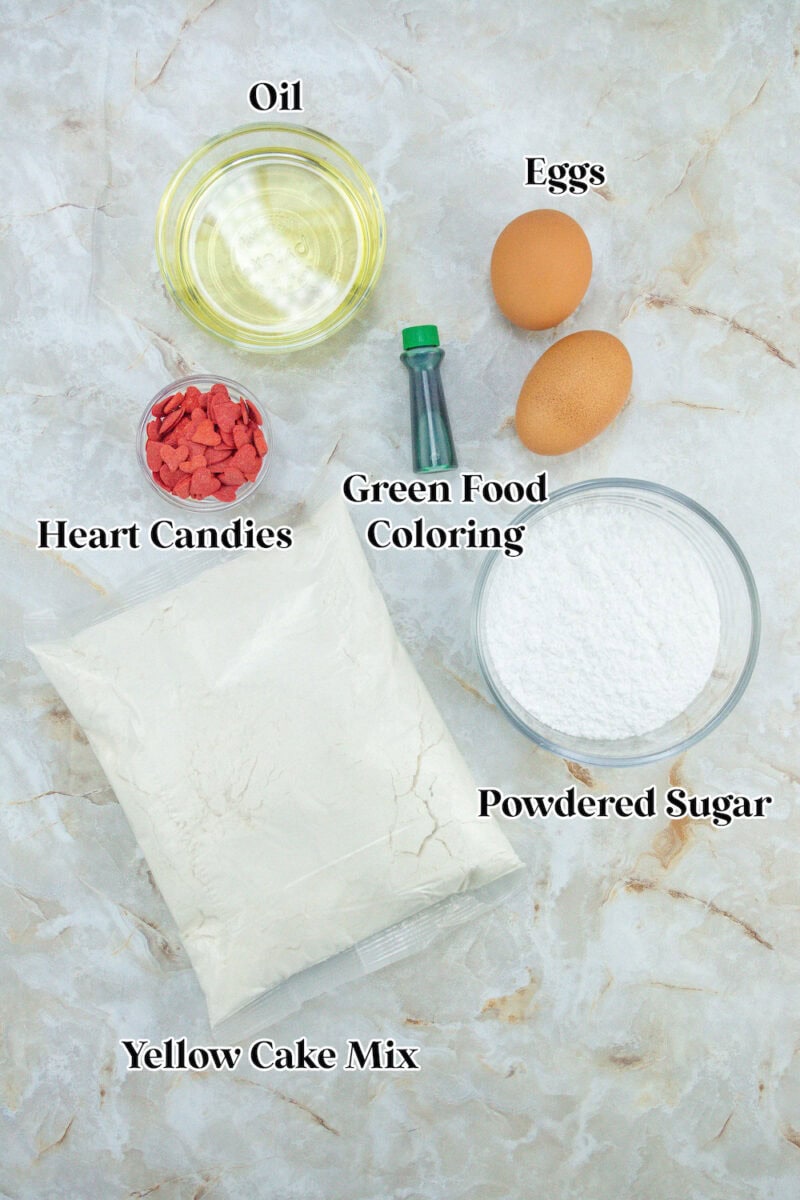 A flat lay of ingredients including oil, eggs, powdered sugar, heart candies, green food coloring, and yellow cake mix on a marble surface.