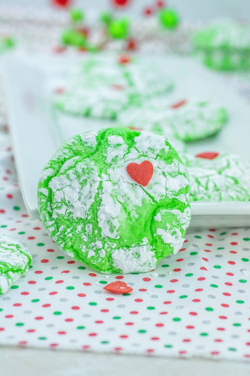 Green crinkle cookies with powdered sugar and red heart-shaped decorations on a polka dot cloth.