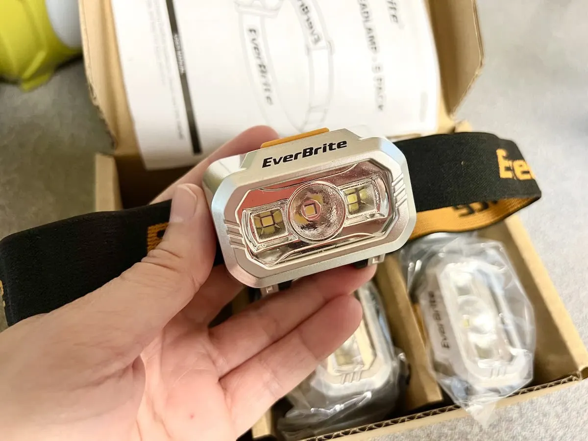 Hand holding an EverBrite headlamp, with multiple headlamps visible in a cardboard box in the background.