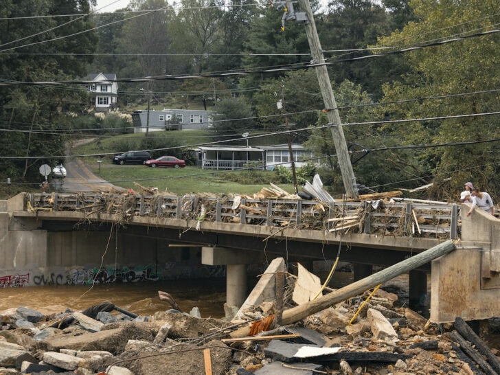 A damaged bridge with debris, fallen utility pole, and wreckage. A person stands near the edge. Residential buildings and parked cars are visible in the background.