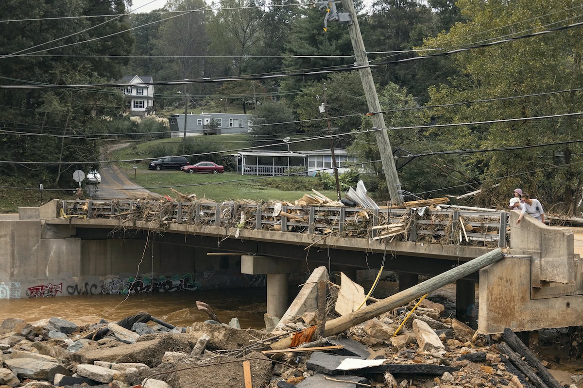 A damaged bridge with debris, fallen utility pole, and wreckage. A person stands near the edge. Residential buildings and parked cars are visible in the background.