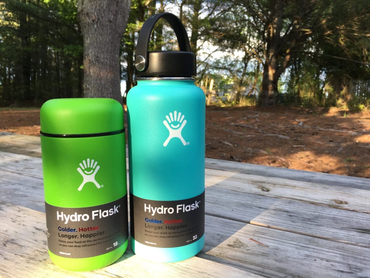 A green Hydro Flask food container and a blue water bottle on a wooden picnic table in a forest setting.