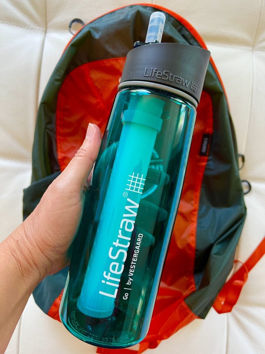 A hand holding a LifeStraw water bottle with a built-in filter, in front of a red and gray backpack.