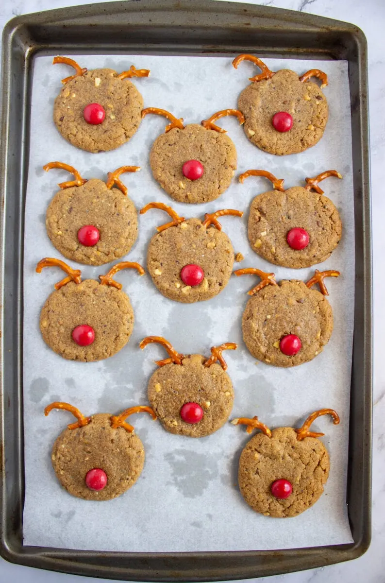 Twelve reindeer-shaped cookies with pretzel antlers and red candy noses on a baking tray lined with parchment paper.