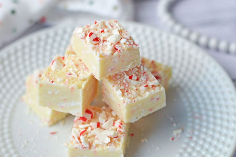 A stack of white chocolate fudge pieces topped with crushed peppermint is displayed on a white plate.
