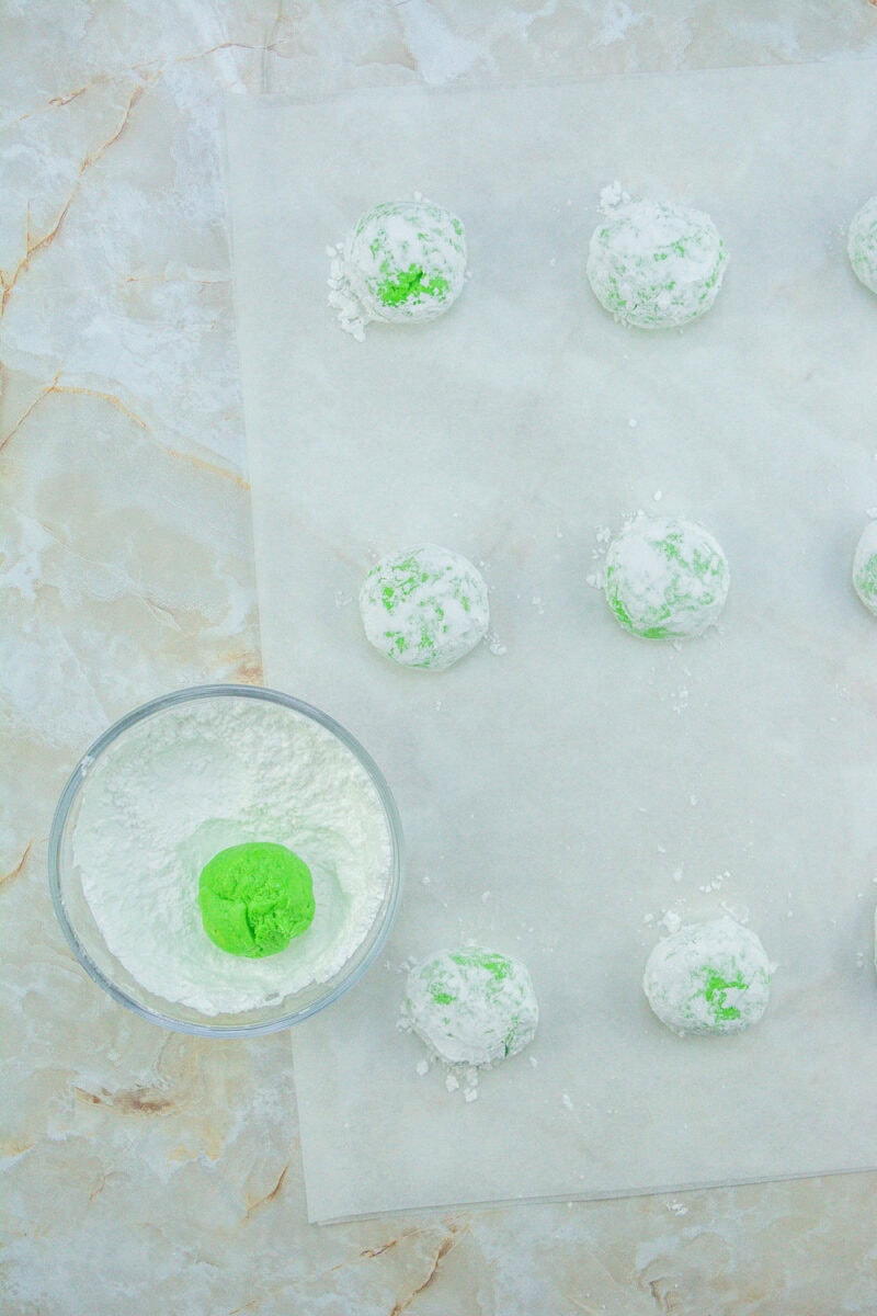 Green cookie dough balls are coated in powdered sugar on parchment paper. A bowl with more dough and sugar is on the side.