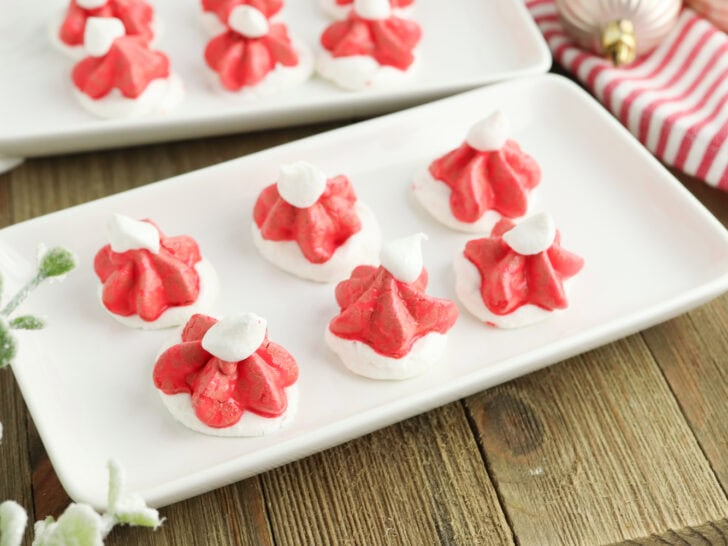 Red and white meringue cookies shaped like Santa hats on a rectangular white plate, placed on a wooden table.