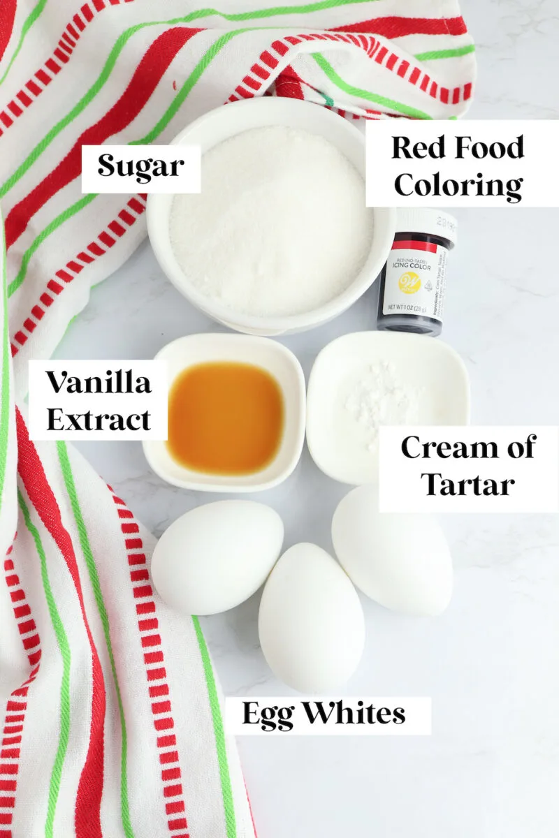 Ingredients for meringue: sugar, red food coloring, vanilla extract, cream of tartar, and three egg whites on a red striped cloth.