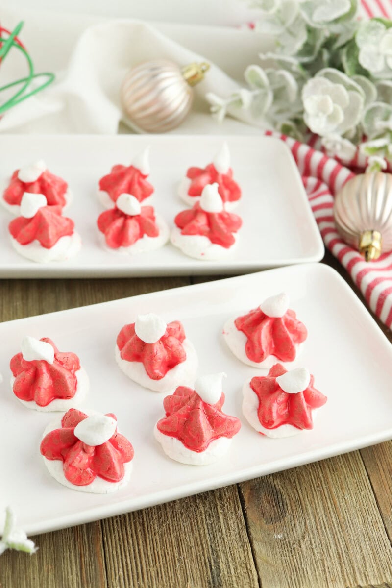 Two plates of festive red and white meringue cookies are displayed on a wooden table, surrounded by Christmas ornaments and greenery.