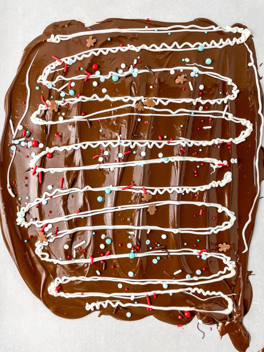 Chocolate bark with white icing drizzles, red and blue sprinkles, and snowflake decorations on a flat surface.