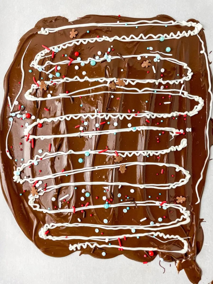 Chocolate bark with white icing drizzles, red and blue sprinkles, and snowflake decorations on a flat surface.