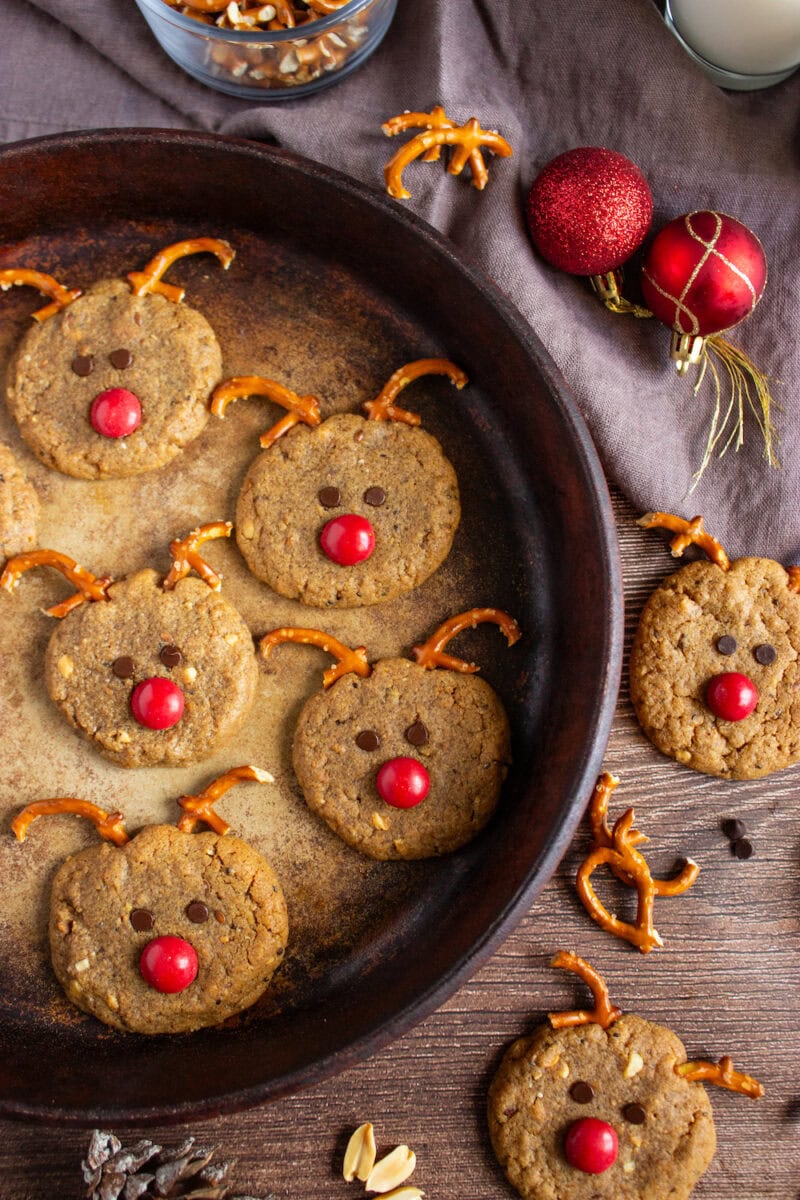 Cookies arranged in a pan, decorated to resemble reindeer faces with pretzel antlers, chocolate eyes, and red candy noses. Surrounding decor includes red ornaments and mixed nuts.