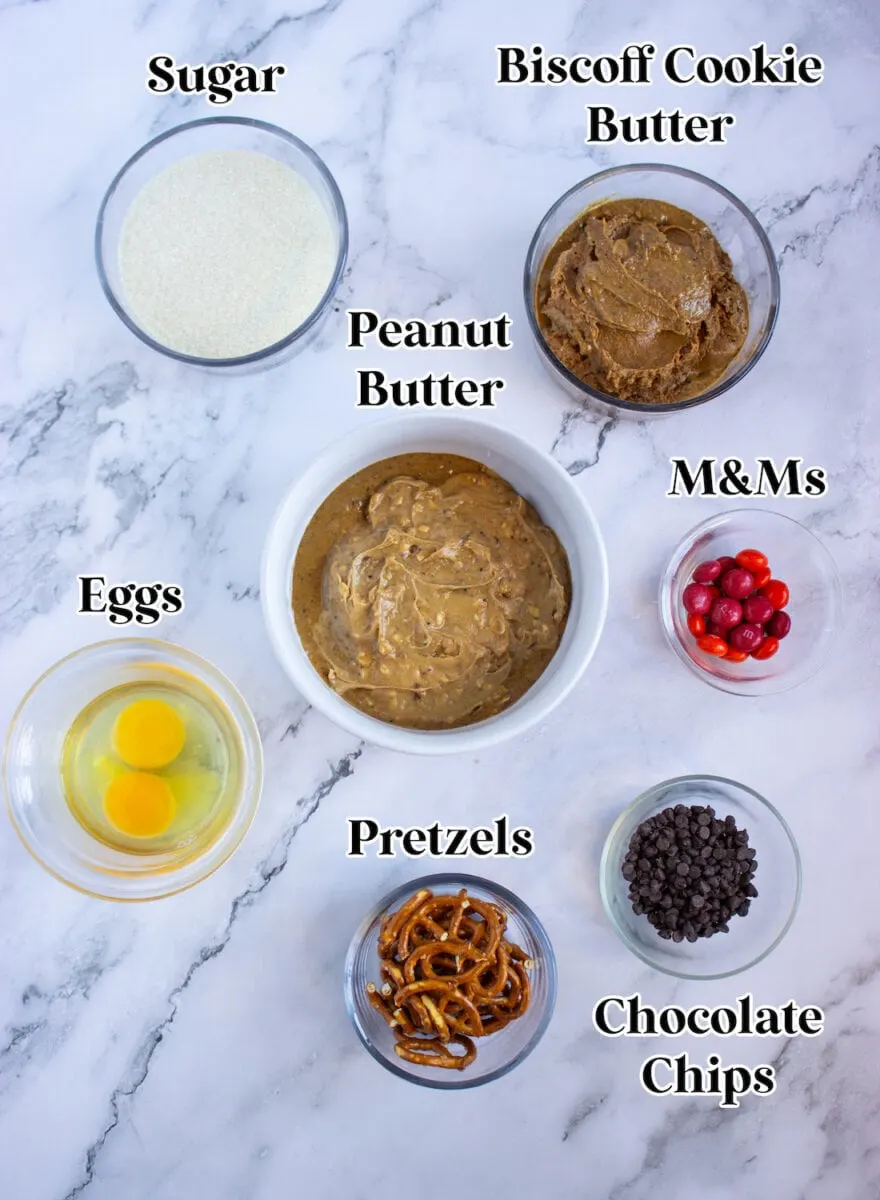 Top-down view of ingredients: sugar, Biscoff cookie butter, peanut butter, M&Ms, eggs, pretzels, and chocolate chips placed in bowls on a marble surface.